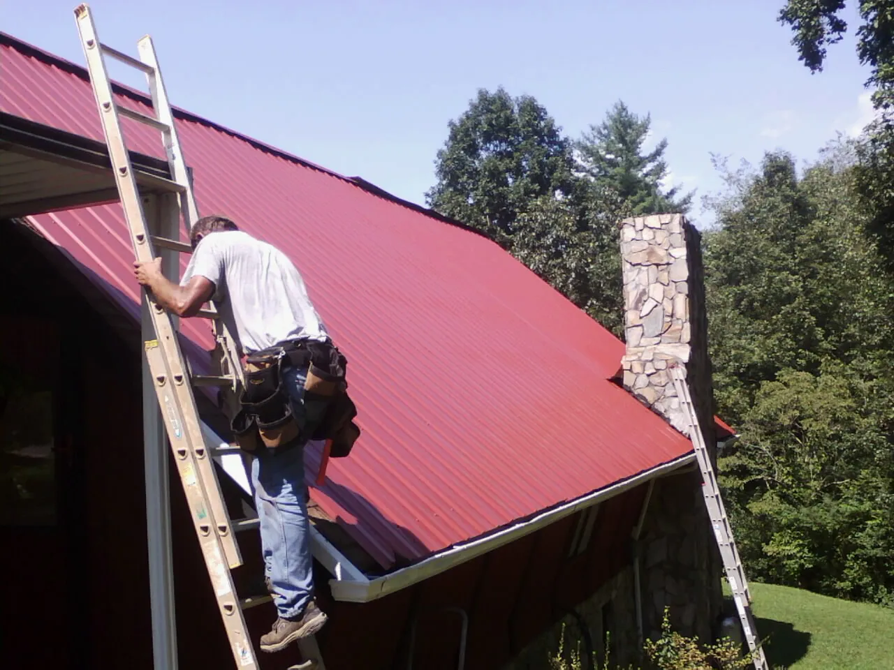 Roofer on a ladder beside a red metal roof and stone chimney — Soffit & Fascia Repair by Roofing Brothers in Circle Pines